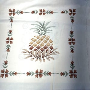 Può includere: Un tessuto bianco con un disegno di ananas marrone e verde. L'ananas è circondato da un bordo di fiori marroni e verdi.