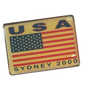 May include: Rectangular pin featuring the American flag with the text "USA" at the top and "SYDNEY 2000" at the bottom. The flag design includes red and yellow stripes with a blue field of white stars. The pin has a gold border.