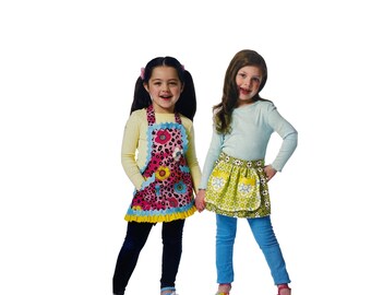 May include: Two girls wearing colorful aprons. One girl is wearing a pink and black floral apron with a blue ruffle trim. The other girl is wearing a green and yellow floral apron with a white ruffle trim. Both girls are smiling and looking at the camera.