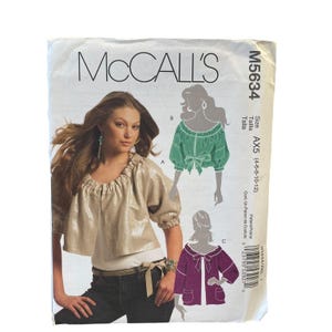 May include: McCall's M5634 sewing pattern envelope featuring a photograph of a model wearing a metallic gold, gathered-neck, short-sleeve top. The envelope includes illustrations of two additional top variations in green and purple, size AX5.