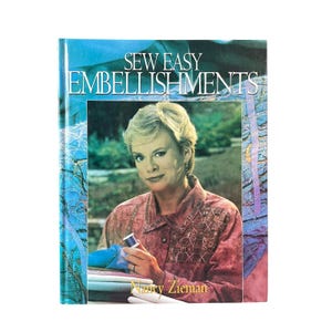 May include: A softcover book titled Sew Easy Embellishments featuring a portrait of a person with blonde hair wearing a textured pink shirt. The cover includes blue and purple abstract patterns and the author name Nancy Zieman in yellow.