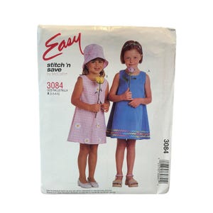Könnte beinhalten: McCall's Easy stitch 'n save Schnittmuster 3084 für Kinderkleider in den Größen 2-3-4-5. Die Packung zeigt zwei Modelle in ärmellosen Kleidern, eines in rosa Vichy-Karo mit Gänseblümchen-Applikationen und Hut, das andere in einfarbigem Blau.