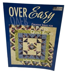 Puede incluir: Un libro de tapa blanda titulado Over Easy: Creative Ideas for Pieced Quilt Backs de Lerlene Nevaril. La cubierta azul oscuro presenta una imagen central de una colcha con patrones florales azules y crema, formas de estrellas geométricas.