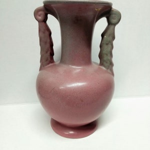 May include: A pink ceramic vase with two handles in the shape of stylized leaves. The vase has a mottled glaze and a rounded body.