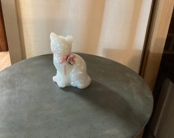 May include: A white iridescent glass cat figurine with a pink bow and flower on its neck. The cat is sitting on a gray surface.