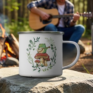 May include: A white enamel mug featuring a printed illustration of a green frog playing a banjo while sitting on a red-capped mushroom. The design is surrounded by a delicate wreath of green leaves and stems, with a silver rim and handle.