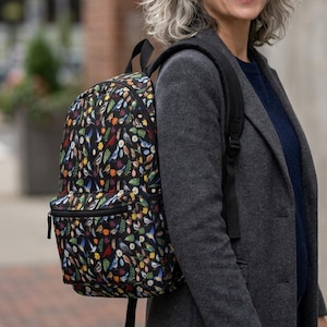 May include: A black backpack featuring a colorful all-over print of various small birds, green leaves, and yellow flowers. The backpack has a front zippered pocket, black adjustable shoulder straps, and a main compartment with a top zipper closure.