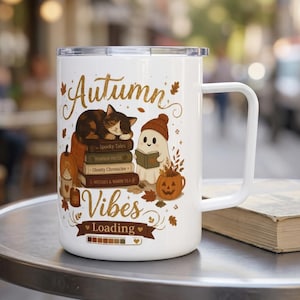 Może przedstawiać: Biały kubek z uchwytem i napisem Autumn Vibes Loading. Grafika przedstawia śpiącego kota na stosie książek, duszka w czapce czytającego książkę, dynię oraz jesienne liście. Na dole znajduje się pasek ładowania.