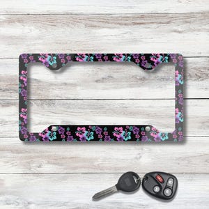 May include: Black license plate frame with a floral pattern of pink, purple, and turquoise hibiscus flowers. The frame is rectangular with a clear center and is set against a white wood background. A car key and remote are in the foreground.