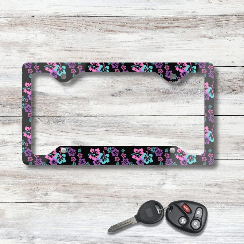 May include: Black license plate frame with a floral pattern of pink, purple, and turquoise hibiscus flowers. The frame is rectangular with a clear center and is set against a white wood background. A car key and remote are in the foreground.