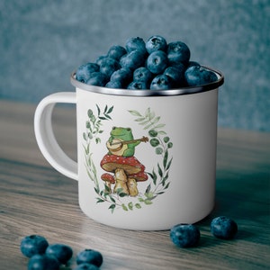 May include: A white enamel mug filled with fresh blueberries. The mug features a printed illustration of a green frog playing a banjo while sitting on a red and white spotted mushroom, surrounded by a delicate green leafy wreath design.