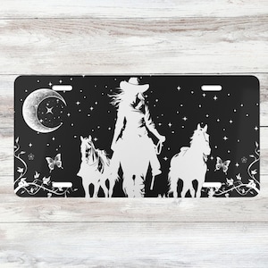 Puede incluir: Silueta en blanco y negro de una vaquera montando a caballo con otros dos caballos a su lado. El fondo es un cielo nocturno negro con una luna creciente y estrellas.