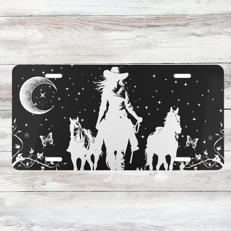 Puede incluir: Silueta en blanco y negro de una vaquera montando a caballo con otros dos caballos a su lado. El fondo es un cielo nocturno negro con una luna creciente y estrellas.