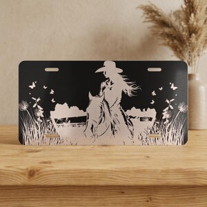 May include: A rectangular metal license plate featuring a black background with a silver-toned silhouette of a person wearing a cowboy hat riding a horse. The design includes tall grasses, wildflowers, and several small butterflies on both sides.