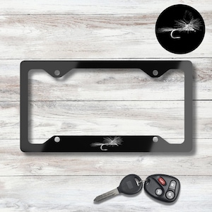 May include: Black license plate frame with a white fly fishing lure design. The frame has a black border and a black background with a white fly fishing lure graphic. A car key and key fob are in the foreground.