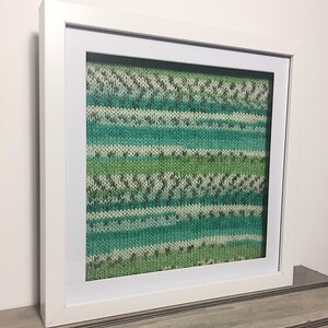 May include: A framed piece of knitted fabric with a striped pattern in shades of green, blue, and white.