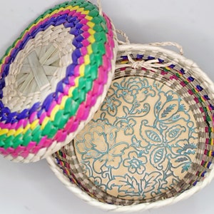 May include: A round woven basket with a lid. The lid is woven with green, pink, yellow, and purple stripes. The inside of the basket is lined with a cream-colored fabric with a blue floral pattern.