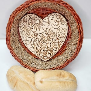 May include: A woven basket with a heart-shaped wooden insert featuring a floral design. The basket has a layered design with brown and tan tones. Two small, baked rolls are in the foreground, suggesting a food-related theme.