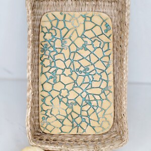 May include: A rectangular, light tan ceramic serving board with a teal geometric design, resting inside a woven basket with a handle. The board features a network of lines and floral shapes. The basket is made of natural-colored woven material.