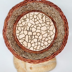 May include: A woven basket with a decorative, light-colored, mosaic-style center. The basket has a layered design with a dark brown outer rim, a tan middle layer, and a light brown inner layer. Two loaves of bread are visible below the basket.