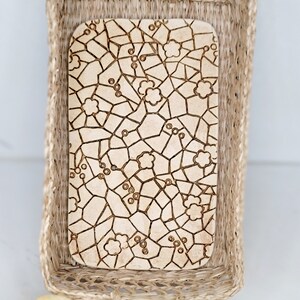 May include: A rectangular wooden serving board with a floral mosaic design sits inside a woven basket. The board's light brown surface features a pattern of interconnected lines and flower-like shapes. The basket is made of natural-colored woven material.
