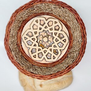 May include: A woven basket with a decorative ceramic plate featuring a geometric design. The basket has a brown rim and a natural fiber interior. Two loaves of bread are visible below the basket.