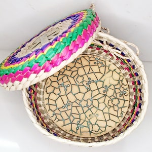 May include: A woven basket with a lid, featuring a colorful design. The lid is adorned with vibrant pink, green, yellow, and blue woven patterns. The basket's interior holds a light-colored, patterned surface with a geometric design.