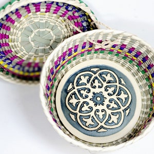 May include: Two woven baskets with colorful accents. The larger basket features a blue and white ceramic base with a floral design. The smaller basket is partially visible above, showcasing a woven pattern with green, purple, and yellow hues.