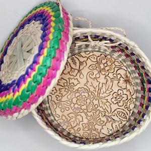 May include: A round woven basket with a lid. The lid is woven with colorful stripes of pink, green, yellow, and purple. The inside of the basket is lined with a cream-colored fabric with a floral pattern.