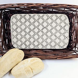 May include: A brown wicker bread basket with a white ceramic insert featuring a geometric pattern. Two white bread rolls are in front of the basket.