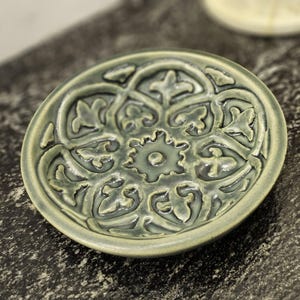May include: A small, green ceramic dish with an intricate, embossed floral design. The dish has a slightly raised rim and a smooth, glazed finish.