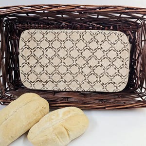 May include: A rectangular woven basket with a decorative patterned insert, and two loaves of bread. The basket is dark brown, and the insert is beige with a repeating geometric design. The bread is light brown.