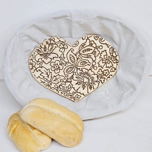 May include: A heart-shaped wooden bread basket with a floral design. The basket is made of natural wood and has a light brown color. The basket is sitting on a white linen cloth.
