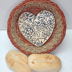 May include: A woven basket with a heart-shaped wooden insert featuring a blue floral design. Two small, golden-brown bread rolls are in the foreground. The basket has a circular shape with multiple layers of woven material.