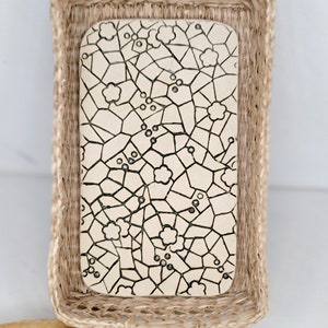 May include: A rectangular serving board with a floral mosaic design sits inside a woven basket. The board has a cream-colored surface with black lines and small flower shapes. The basket is made of natural woven fibers.