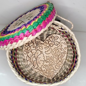 May include: A woven basket with a lid, featuring a heart-shaped wooden insert. The lid and the basket's rim are adorned with vibrant, multicolored woven patterns. The heart-shaped insert is decorated with a floral design.