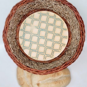 May include: A woven wicker basket with a round, wooden base featuring a geometric pattern in shades of blue and beige. The basket is sitting on top of two loaves of bread.