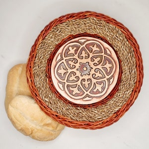 May include: A round woven coaster with a brown and tan design. The coaster has a center piece with a brown and red geometric pattern.