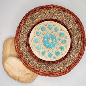 May include: A woven placemat with a brown rim and a natural fiber center, topped with a ceramic coaster featuring a turquoise geometric design. Two loaves of bread are on the left.