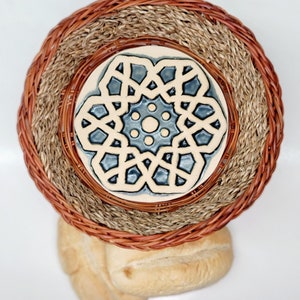 May include: A woven basket with a decorative ceramic tile featuring a blue and white geometric pattern. The basket has a brown rim and a natural fiber interior. Two rolls are visible below the basket.
