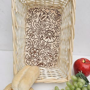 May include: A white wicker bread basket with a floral patterned liner. The basket is filled with two loaves of bread, a bunch of green grapes, and a red apple.