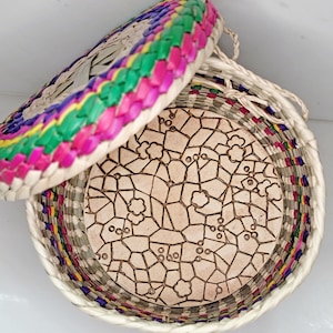 May include: A round, woven basket with a lid. The lid is decorated with a rainbow of colors, including pink, green, and yellow. The basket's interior features a light brown, patterned design. The basket is made of natural materials.