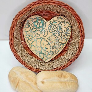 May include: A heart-shaped decorative piece with teal floral designs sits inside a woven basket. Two small, golden-brown bread rolls are in front of the basket. The basket has a brown and tan color scheme.