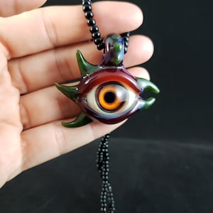 Puede incluir: Un collar con un colgante de ojo de vidrio con un ojo rojo, naranja y amarillo, rodeado de vidrio verde y morado. El collar está hecho de cuentas negras.