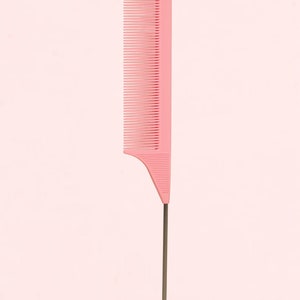 May include: A pink plastic comb with a long, thin metal tail. The comb has fine teeth and a handle with a hook. The background is a solid light pink.