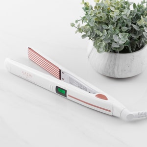 Może przedstawiać: Biała i różowo-złota prostownica do włosów z nazwą marki "AASHI" wydrukowaną z boku. Prostownica posiada cyfrowy wyświetlacz i różowo-złoty akcent. Płytki mają żebrowany wzór z różowo-złotymi paskami. Mała roślina doniczkowa znajduje się w tle.