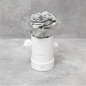 Puede incluir: Una rosa plateada en una caja redonda blanca con una cinta blanca. La caja tiene la inscripci&oacute;n dorada "SAAYA ROSE". La rosa met&aacute;lica es el elemento central, su color contrasta con el blanco de la caja y la cinta.