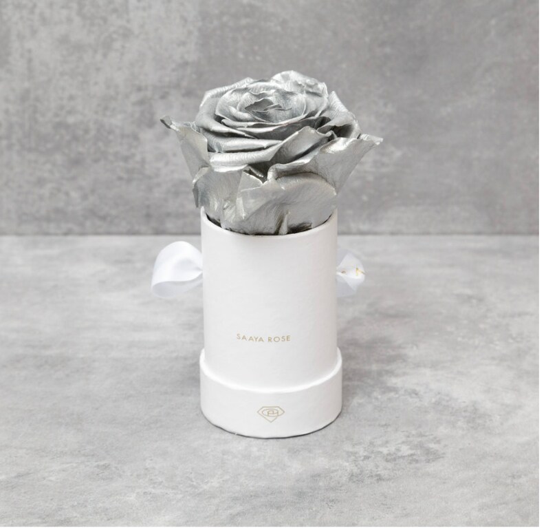 Puede incluir: Una rosa plateada en una caja redonda blanca con una cinta blanca. La caja tiene la inscripci&oacute;n dorada "SAAYA ROSE". La rosa met&aacute;lica es el elemento central, su color contrasta con el blanco de la caja y la cinta.