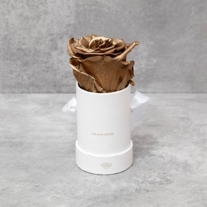 Puede incluir: Una rosa dorada en una caja redonda blanca con el texto "SAAYA ROSE" impreso en la parte delantera. La caja est&aacute; atada con una cinta blanca. La rosa es el punto focal, con su acabado met&aacute;lico que contrasta con la caja blanca.