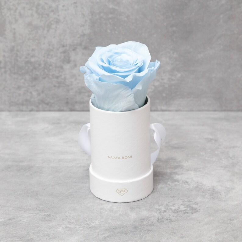 Puede incluir: Una rosa azul claro en una caja de regalo cil&iacute;ndrica blanca. La caja tiene una tapa a juego, una cinta blanca y el texto dorado "SAAYA ROSE". La rosa es el foco principal, con sus delicados p&eacute;talos y su color suave. La caja mide unos 8 cm de alto.